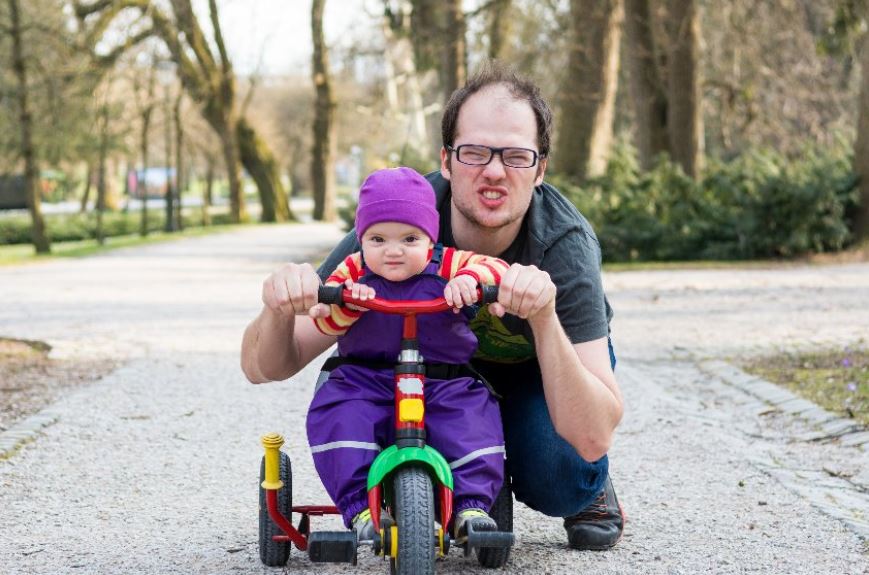 un enfant qui fait du tricycle sur le trottoir avec son pere a ses cotes