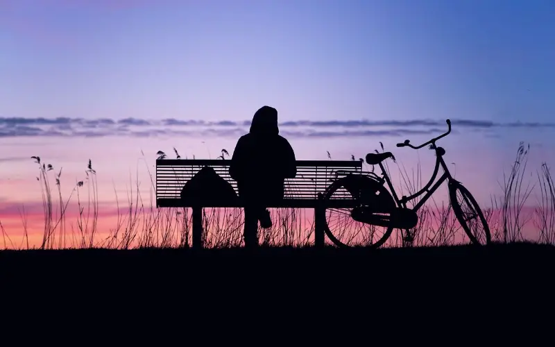 une personne assise sur un banc avec son velo a cote au couche du soleil